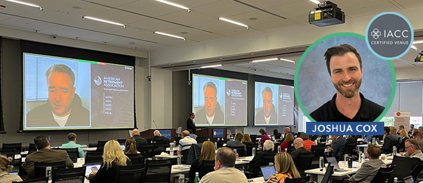 OLC auditorium full of attendees with the main presenter coming in virtually on 5 large screens with his presentation next to him on the screen. Joshua Cox, AV Media Production Manager is shown as an inset image -- he makes the magic happen